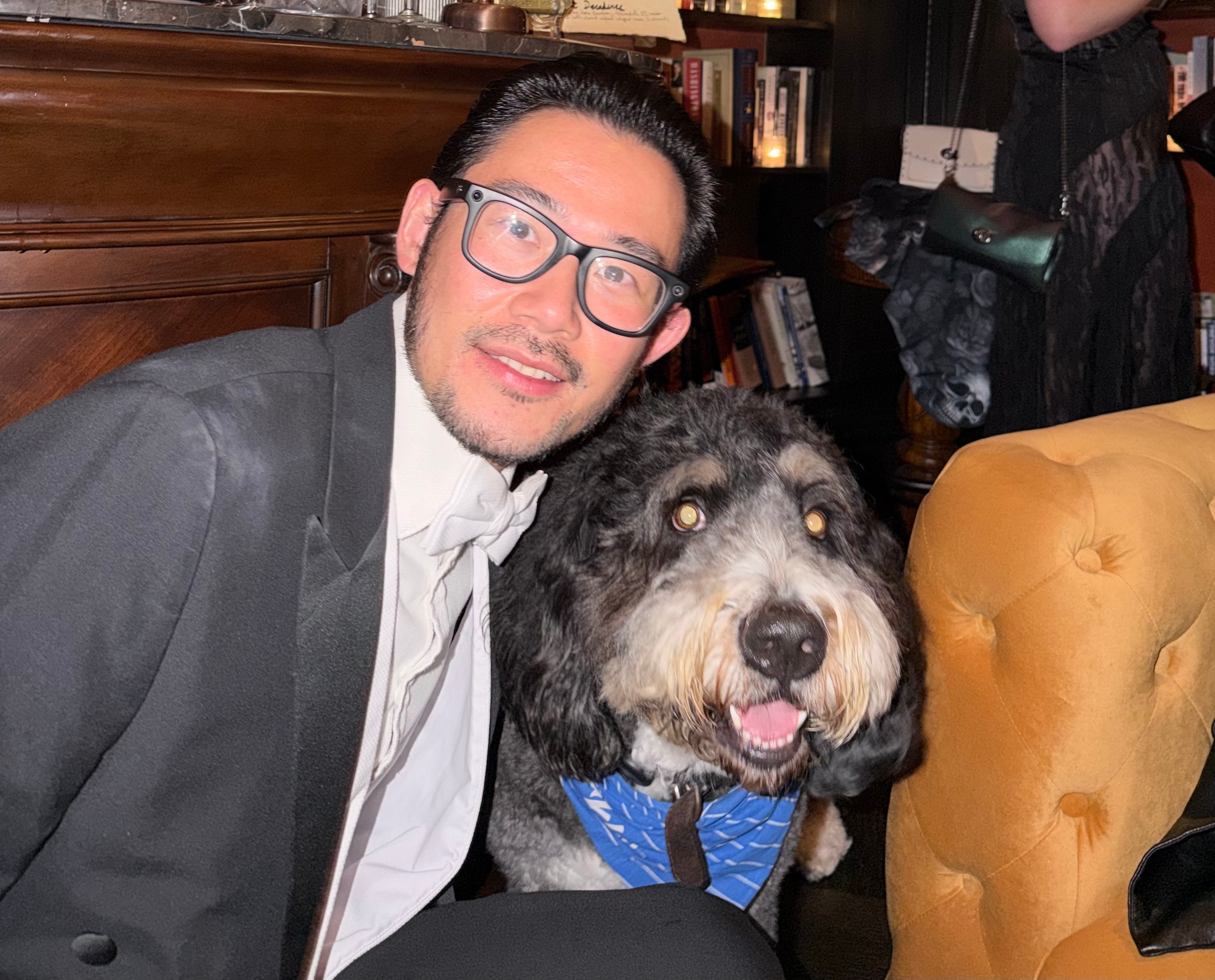 Jay crouching next to a fluffy black, gray and white doodle wearing a blue bandana in a bookshelf-lined bar