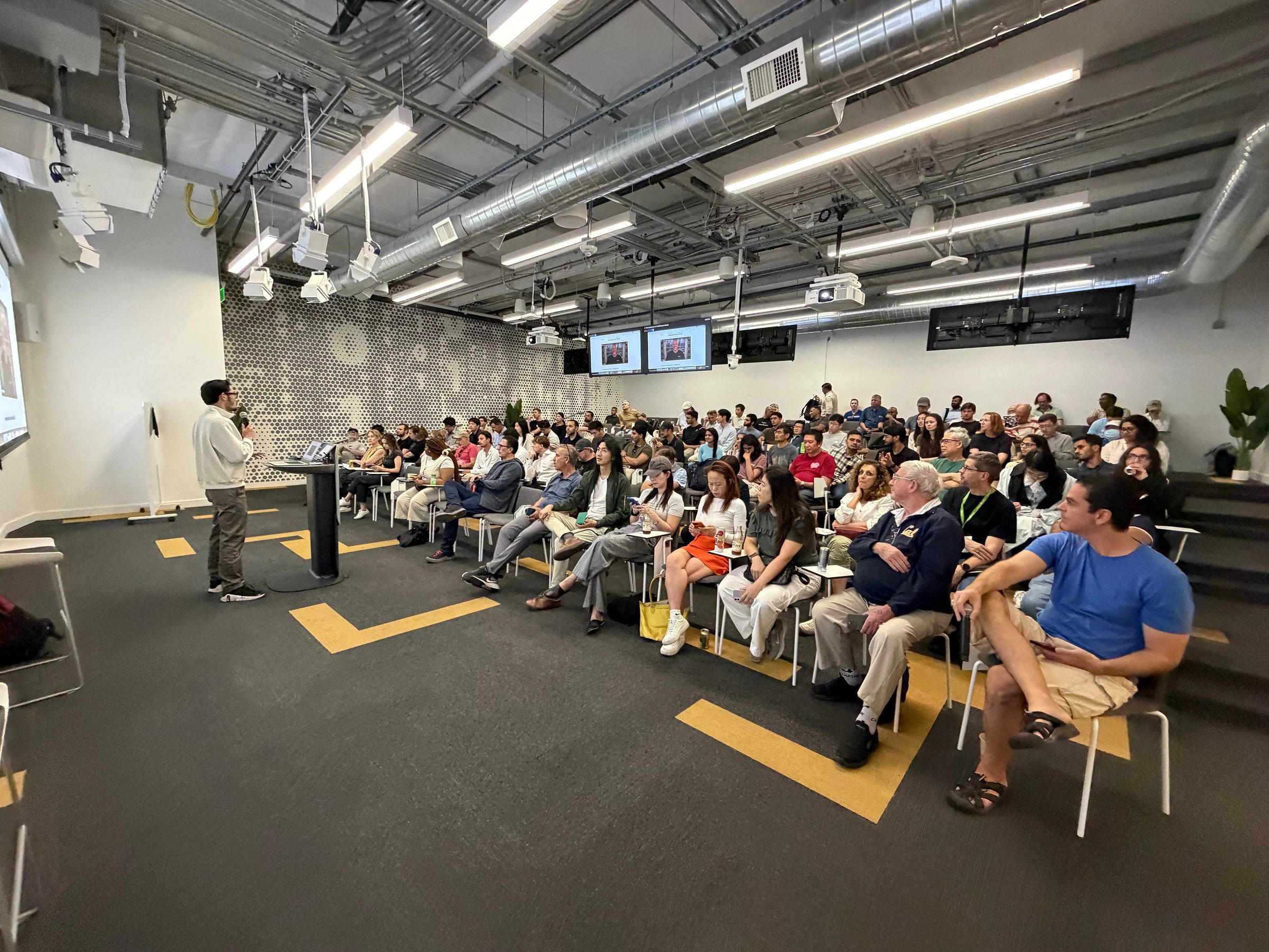 Wide view of a packed meetup audience listening to a speaker at a podium